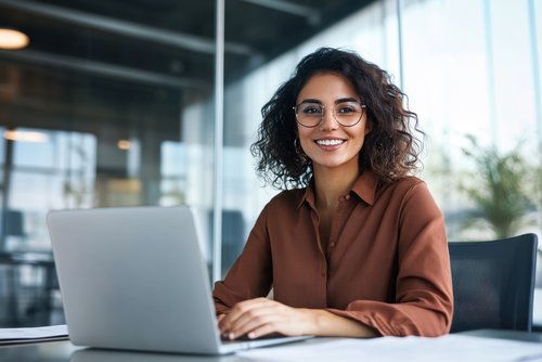 Eine Frau mit lockigem Haar und Brille sitzt an einem Schreibtisch und lächelt, während sie an einem Laptop arbeitet.