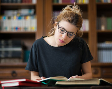 Karriere-Psychologisches Coaching Eine junge Frau mit Brille liest konzentriert in einem Buch, während sie an einem Tisch in einer Bibliothek sitzt.
