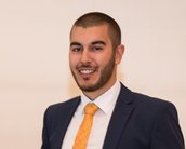 Ein Mann mit Bart trägt einen Anzug und eine orangefarbene Krawatte. Er lächelt und steht vor einem neutralen Hintergrund.