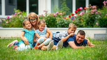 Eine Frau und vier Kinder liegen lächelnd auf einer Wiese, umgeben von bunten Blumen. Die Kinder sind in verschiedenen Outfits gekleidet und scheinen Spaß zu haben.