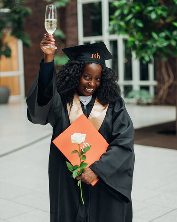 A graduate in a black gown and cap holds a glass of champagne in one hand and a diploma with a rose in the other.