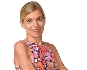 Eine Frau mit blonden Haaren trägt ein blumiges, ärmelloses Kleid und steht mit verschränkten Armen vor einem weißen Hintergrund.