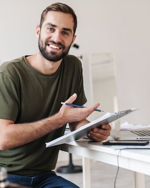 Ein lächelnder Mann sitzt an einem Tisch, hält einen Stift und eine Mappe, während er vor einem Laptop arbeitet. Pflanzen sind im Hintergrund sichtbar.