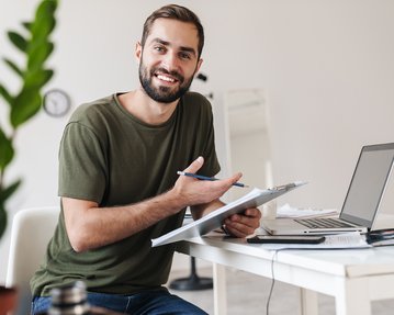 Ein lächelnder Mann sitzt an einem Tisch, hält einen Stift und eine Mappe, während er vor einem Laptop arbeitet. Pflanzen sind im Hintergrund sichtbar.