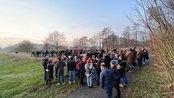 Eine große Menschenmenge versammelt sich auf einem Weg in einer parkähnlichen Umgebung, um ein Ereignis zu beobachten.