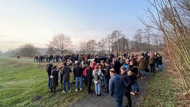 Eine große Menschenmenge versammelt sich auf einem Weg in einer parkähnlichen Umgebung, um ein Ereignis zu beobachten.