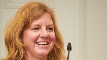 Eine lächelnde Frau mit lockigem, schulterlangem, blondem Haar trägt ein grünes Oberteil. Sie steht vor einem Mikrofon und scheint an einem öffentlichen Event oder einer Präsentation teilzunehmen.