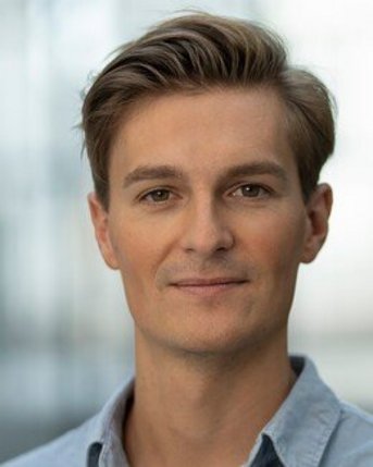 Ein junger Mann mit kurzen, gestylten Haaren und einem hellblauen Hemd schaut direkt in die Kamera. Der Hintergrund ist unscharf.