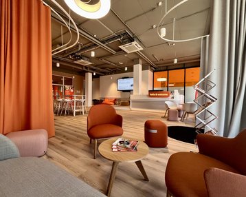 Moderne Lounge mit gemütlichen Sesseln, einem runden Tisch und einem Empfangsbereich im Hintergrund. Große Fenster und warme Farben dominieren den Raum.