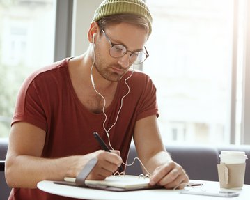 Ein Mann mit Brille und Kopfhörern schreibt in ein Notizbuch, während er an einem Tisch sitzt. Neben ihm steht eine Kaffeetasse.
