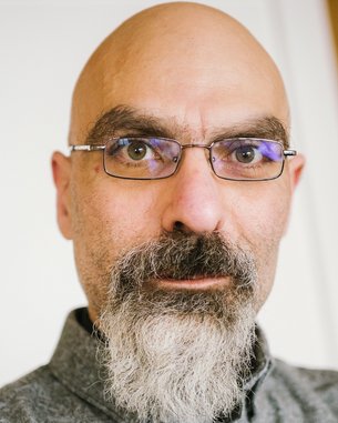 A man with a bald head and glasses, who has a gray beard. He is looking directly at the camera and wearing a gray shirt. The background is blurred and bright.