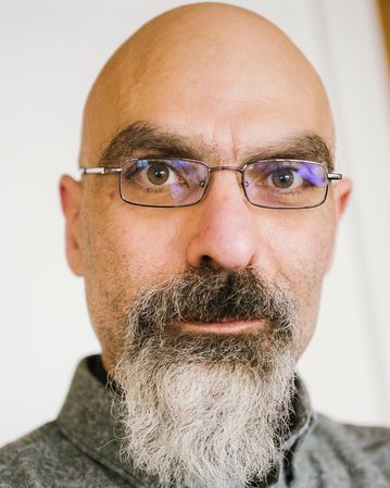 A man with a bald head and glasses, who has a gray beard. He is looking directly at the camera and wearing a gray shirt. The background is blurred and bright.