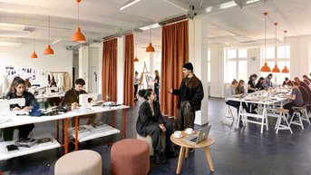 A creative workspace with several people working at tables. Some are discussing while others are working on projects or laptops.