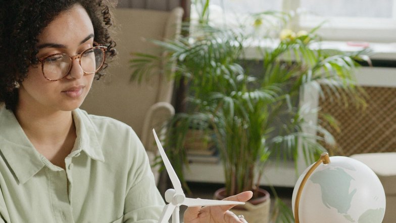 Eine Frau mit lockigem Haar und Brille sitzt an einem Tisch und zeigt auf ein Modell eines Windrads und kleine Häuser, während eine Weltkugel im Hintergrund steht. Pflanzen sind ebenfalls sichtbar.