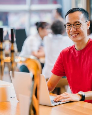 A man with glasses is sitting at a table in a café, working on a laptop. He is wearing a red T-shirt and smiling. In the foreground, a coffee cup and a coffee machine are visible.