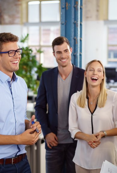 Eine Gruppe von fünf Personen in einem Büro, die lächeln und miteinander sprechen. Ein Mann präsentiert an einer Tafel.