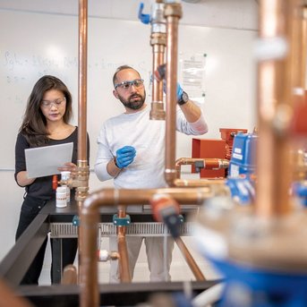 Two people in a laboratory, a woman with notes and a man examining a copper wire. In the background, various equipment and a chalkboard are visible.