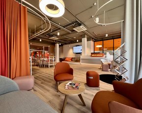 A modern, inviting office with cozy seating, warm colors, and a reception area. Large windows let in plenty of light, while a screen displays information.