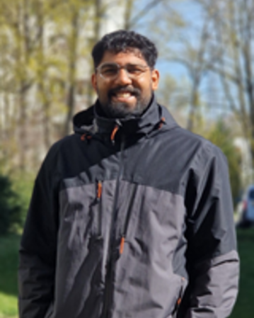 Ein Mann mit Brille trägt eine schwarze Jacke und steht im Freien, umgeben von Bäumen und einem verschwommenen Hintergrund.