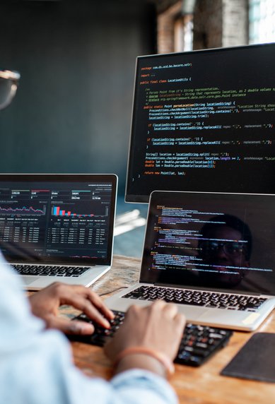 Ein Programmierer arbeitet an mehreren Bildschirmen, auf denen Code und Datenanalysen angezeigt werden. Der Raum hat große Fenster und eine moderne, kreative Atmosphäre.