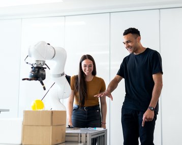Zwei Personen stehen an einem Tisch und beobachten einen Roboterarm, der eine gelbe Kugel greift. Beide wirken interessiert und lächeln, während sie dem Roboter zuschauen.