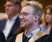 Ein junger Mann mit Brille und blonden Haaren trägt eine Graduierungsrobe und lächelt während einer Zeremonie.
