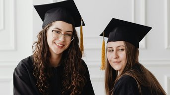 Two graduates in black gowns and caps pose together. One holds a diploma in her hand while the other smiles. The background is simple and bright, drawing attention to the graduates.
