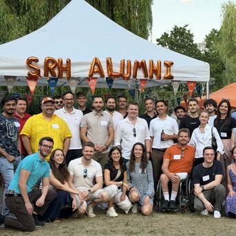 Gruppentreffen von SRH-Alumni unter einem Zelt, mit Teilnehmern in verschiedenen Outfits, lächelnd und stehend, um eine Bannerdekoration.
