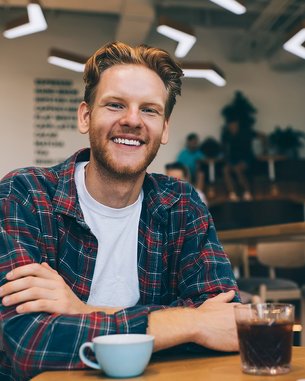 Ein junger Mann mit einem lässigen Outfit sitzt an einem Tisch in einem Café. Vor ihm stehen eine Tasse Kaffee und ein Glas mit einem dunklen Getränk. Er lächelt freundlich in die Kamera.