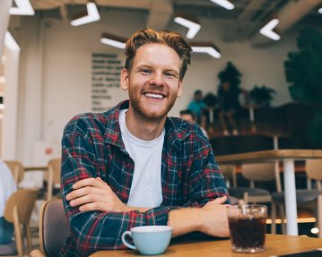 Ein junger Mann mit einem lässigen Outfit sitzt an einem Tisch in einem Café. Vor ihm stehen eine Tasse Kaffee und ein Glas mit einem dunklen Getränk. Er lächelt freundlich in die Kamera.