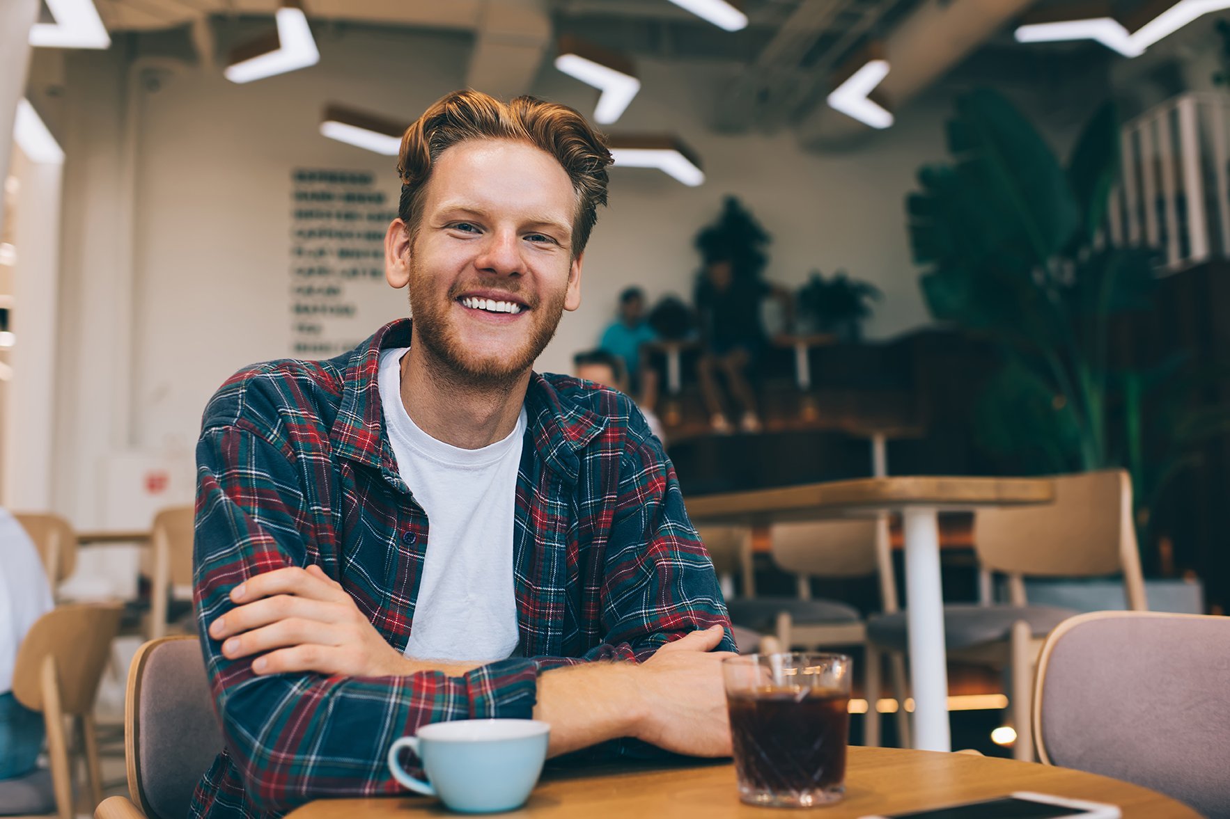 Ein junger Mann mit einem lässigen Outfit sitzt an einem Tisch in einem Café. Vor ihm stehen eine Tasse Kaffee und ein Glas mit einem dunklen Getränk. Er lächelt freundlich in die Kamera.