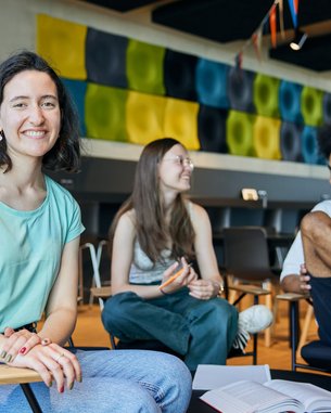 Drei junge Erwachsene sitzen in einem modernen Raum. Eine Frau mit kurzen, lockigen Haaren lächelt in die Kamera, während zwei andere Personen, eine Frau und ein Mann, miteinander sprechen. Bunte Wanddekorationen sind im Hintergrund sichtbar.