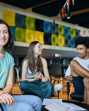 Drei junge Erwachsene sitzen in einem modernen Raum. Eine Frau mit kurzen, lockigen Haaren lächelt in die Kamera, während zwei andere Personen, eine Frau und ein Mann, miteinander sprechen. Bunte Wanddekorationen sind im Hintergrund sichtbar.