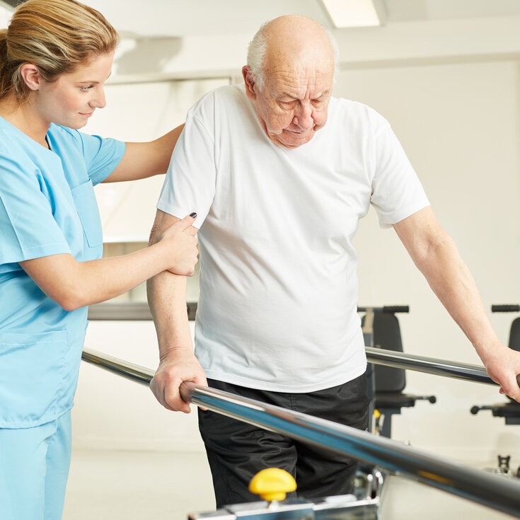Eine Krankenschwester unterstützt einen älteren Mann beim Gehen an einem Haltegriff in einer Rehabilitationsumgebung.
