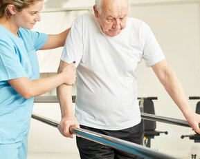 Eine Krankenschwester unterstützt einen älteren Mann beim Gehen an einem Haltegriff in einer Rehabilitationsumgebung.