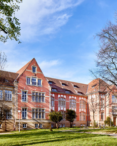 Ein historisches Gebäude mit roten Ziegeln, umgeben von Bäumen und Rasenflächen, unter einem klaren blauen Himmel. Die Architektur zeigt große Fenster und einen einladenden Eingang.