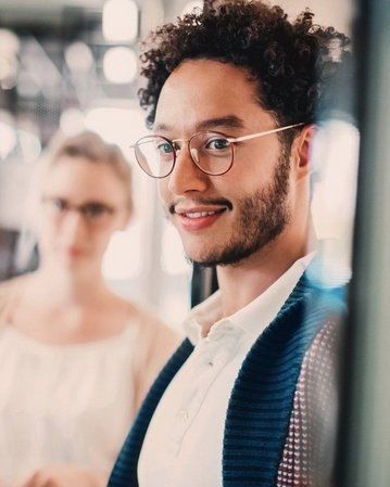 A young man with curly hair and glasses smiles at the camera, while a woman stands in the background, also looking on. They are in a modern, bright room.