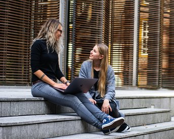 Zwei Frauen sitzen auf Treppenstufen und unterhalten sich. Eine von ihnen hat einen Laptop auf dem Schoß, während die andere aufmerksam zuhört. Im Hintergrund sind moderne Holzlatten zu sehen.