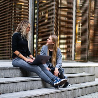 Zwei Frauen sitzen auf Treppenstufen und unterhalten sich. Eine von ihnen hat einen Laptop auf dem Schoß, während die andere aufmerksam zuhört. Im Hintergrund sind moderne Holzlatten zu sehen.