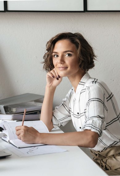 A woman sits at a desk, smiling and holding a pencil. On the table are notes and a laptop. In the background, plants and artworks are visible.