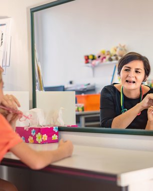 Eine Frau erklärt einem Mädchen im orangefarbenen Shirt etwas vor einem Spiegel. Das Mädchen hält eine Zahnbürste in der Hand. Auf dem Tisch steht eine Box mit Taschentüchern und Spielzeug im Hintergrund.