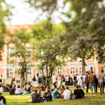 People are gathered in a meadow, some sitting and others standing, surrounded by trees and a historic building in the background.