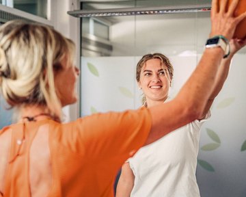 A person is holding a ball up while another person watches in a training room. Both are wearing athletic clothing. The room has a bright, cheerful atmosphere with plant motifs in the background.