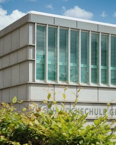 Modernes Schulgebäude mit großen Fenstern und einer klaren, geometrischen Fassade, umgeben von Bäumen und Sträuchern.