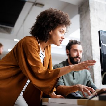 A woman with curly hair is explaining something to a man at a computer in a modern office. In the background, other people can be seen.