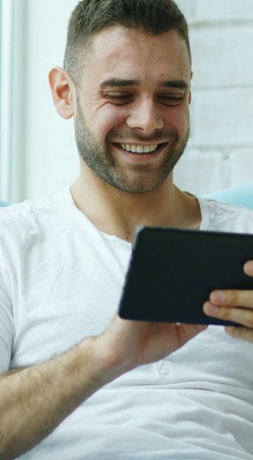 Ein Mann sitzt in einem blauen Sessel und schaut lächelnd auf ein Tablet. Helle Fenster und eine Ziegelwand sind im Hintergrund sichtbar.