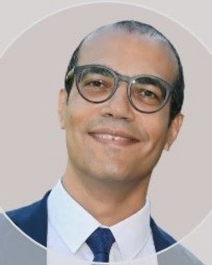 Ein Mann mit Brille und kurzem, dunklem Haar lächelt in die Kamera. Er trägt ein weißes Hemd und eine dunkle Krawatte.