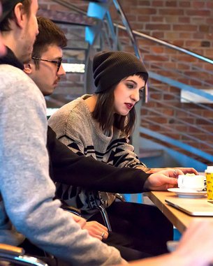 A group of four people is sitting at a table with laptops. One person in the middle, wearing a hat, is looking at the screen and interacting with a laptop. There are cups and a yellow mug on the table.