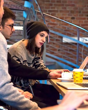 Eine Gruppe von vier Personen sitzt an einem Tisch mit Laptops. Eine Person in der Mitte, mit einer Mütze, betrachtet den Bildschirm und interagiert mit einem Laptop. Auf dem Tisch stehen Tassen und ein gelbes Becher.