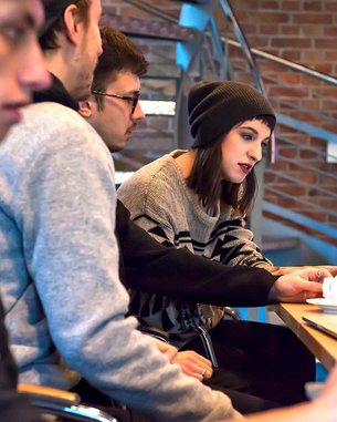 A group of four people is sitting at a table with laptops. One person in the middle, wearing a hat, is looking at the screen and interacting with a laptop. There are cups and a yellow mug on the table.